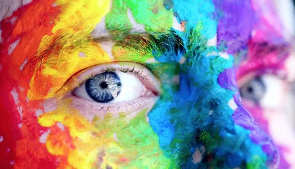 Wir sehen den Ausschnitt eines jungen Gesichtes, das wild in Regenbogenfarben geschminkt ist. Nur die Augen sind frei zu sehen