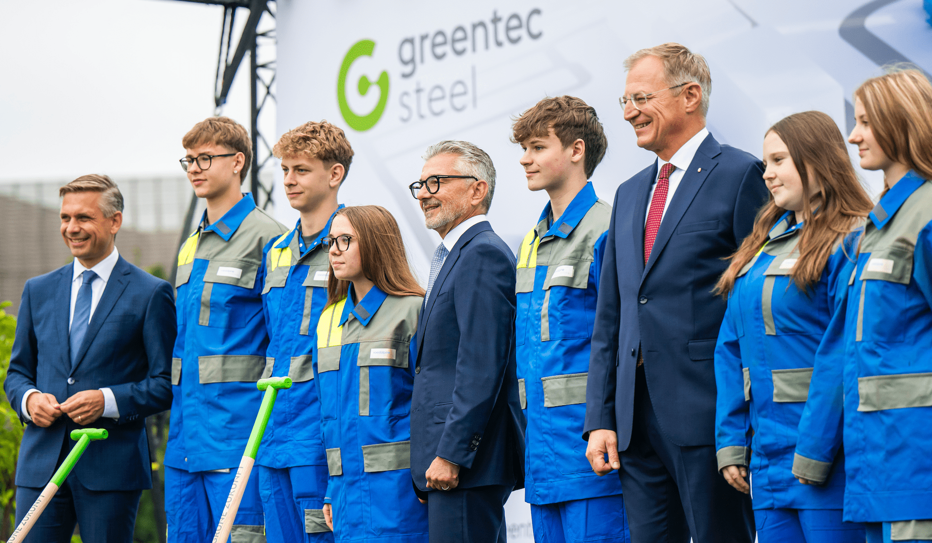 Gruppenfoto von Jugendlichen in blauen Arbeitsanzügen mit zwei Männern in Anzügen vor einem Banner mit dem Schriftzug greentec steel.