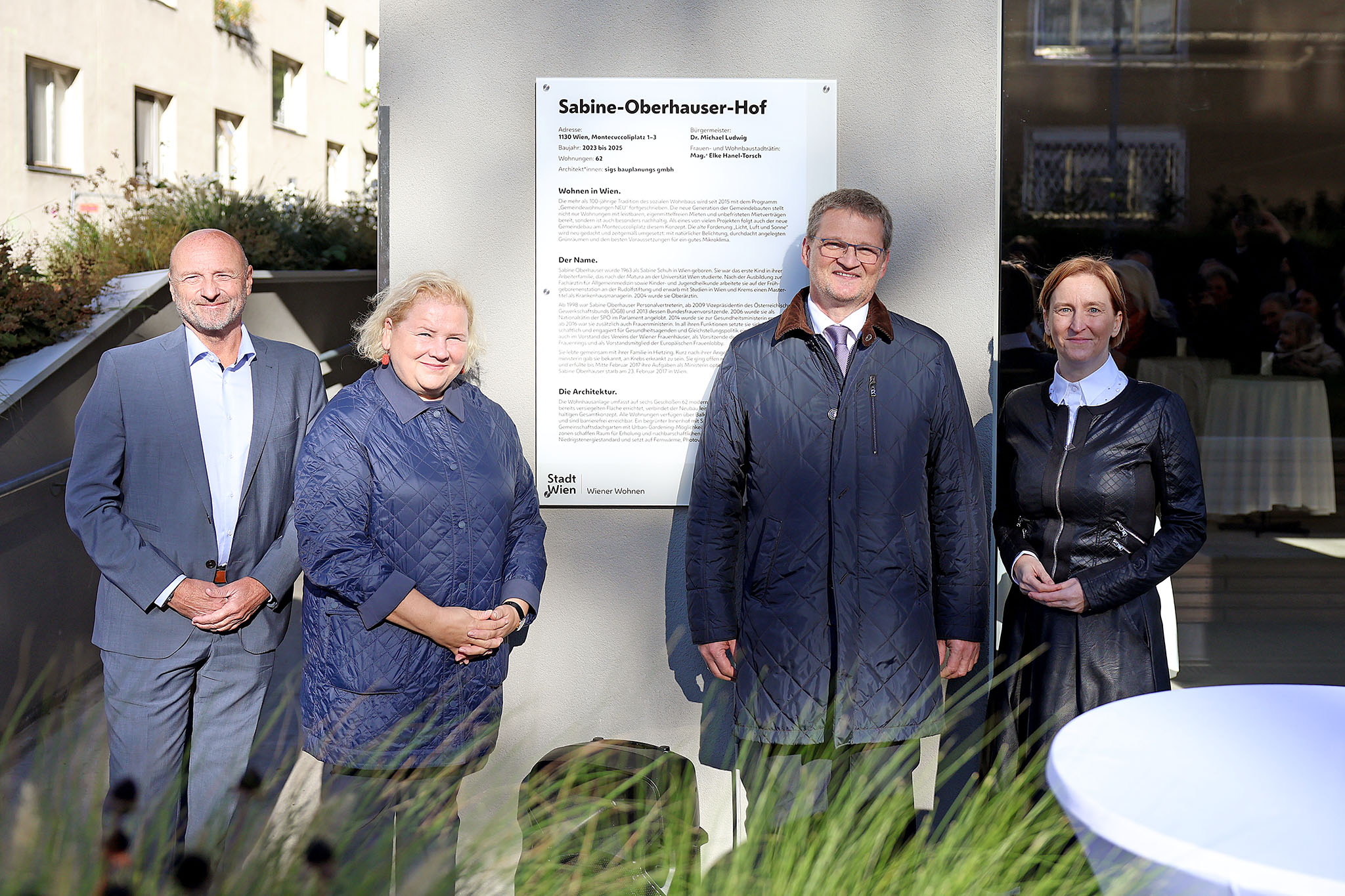 Vier Personen stehen vor einer Informationstafel zum Sabine-Oberhauser-Hof in Wien bei sonnigem Wetter.