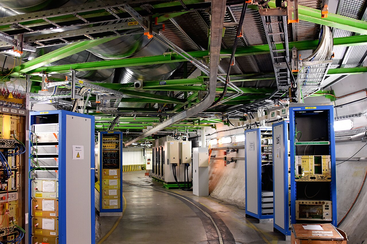 Blick in einen technischen Gang mit Server- und Steuerungsschränken, Kabeltrassen an der Decke und Beleuchtung in einem Forschungslabor.