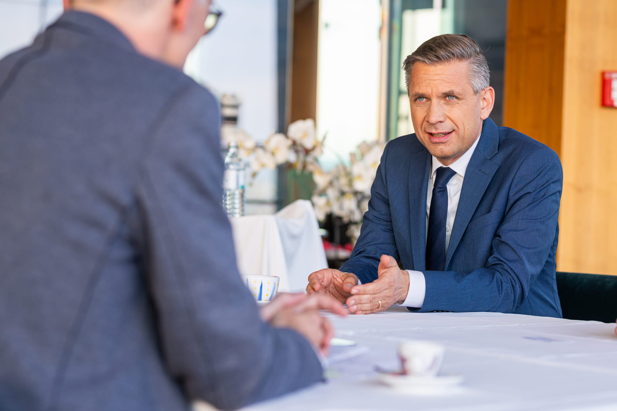 Zwei Männer im Anzug sitzen an einem Tisch, einer spricht gestikulierend, im Hintergrund Blumen und Fenster mit Tageslicht.